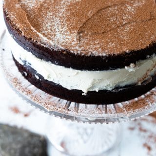 A chocolate cake with white frosting on a glass cake plate with a silver cake knife next to it.