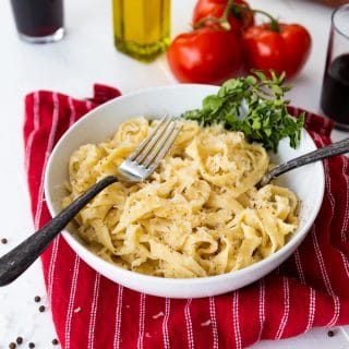 Pasta noodles in a white bowl with two forks and fresh herbs in the bowl, on a red striped napkin with whole tomatoes and two glasses of red wine and a bottle of olive oil in the background