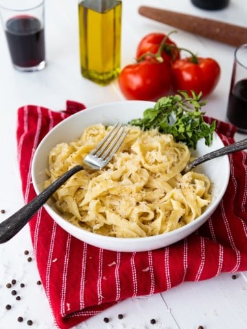 Pasta noodles in a white bowl with two forks and fresh herbs in the bowl, on a red striped napkin with whole tomatoes and two glasses of red wine and a bottle of olive oil in the background