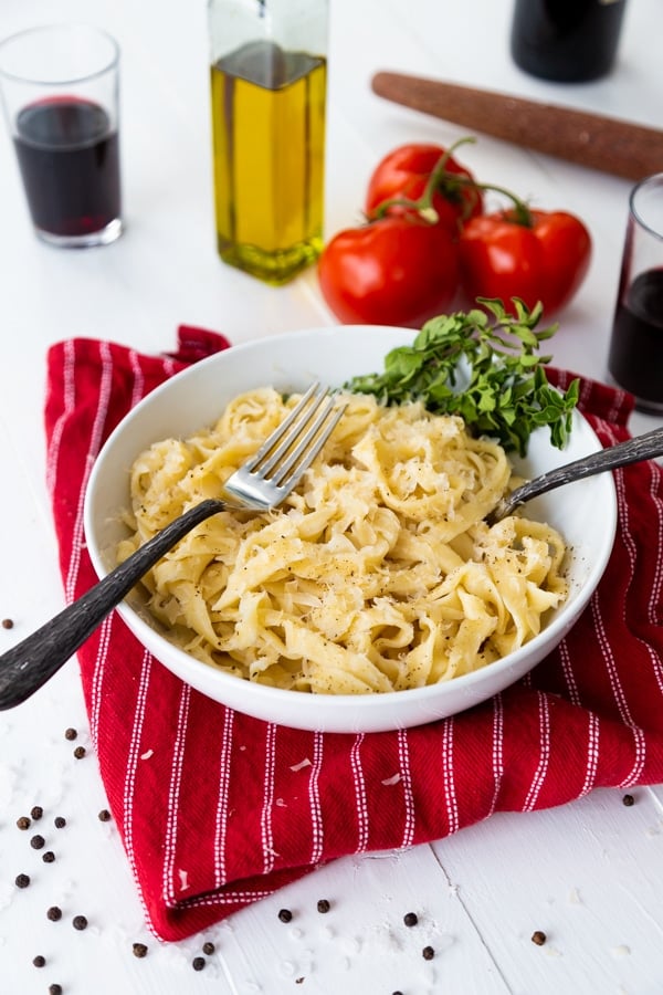 Pasta noodles in a white bowl with two forks and fresh herbs in the bowl, on a red striped napkin with whole tomatoes and two glasses of red wine and a bottle of olive oil in the background