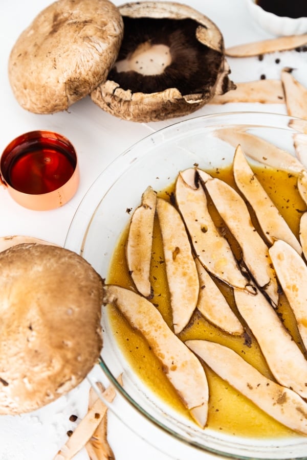 Sliced mushrooms in a glass pie plate marinating in liquid smoke with a cup of maple syrup and mushrooms next to the plate