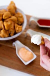 Veganosity's favorite recipe caulflower poppers being dipped in a vegan ranch sauce with a bowl of cauliflower poppers in the background with vegan ranch, vegan sriracha mayo, and vegan hot sauce in three separate white dishes on a wood board.