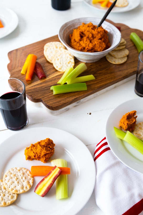 A wood and marble board with a white bowl of curry dip and vegetables and crackers and two white plates with some dip, crackers and vegetables with two glasses of red wine. 