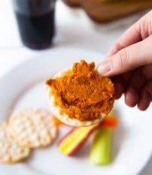 Curry dip on a cracker with a white plate with extra crackers and sliced vegetables.