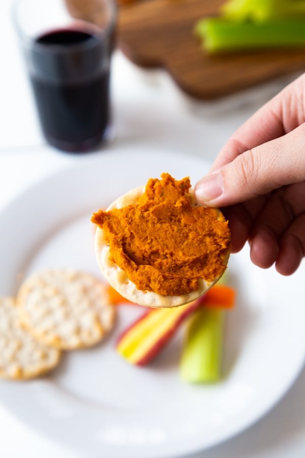 Curry dip on a cracker with a white plate with extra crackers and sliced vegetables.