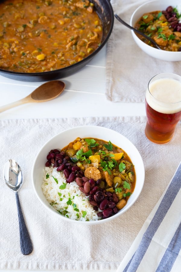 An iron skillet with gumbo and two white bowls with gumbo, rice, and beans with a glass of beer and napkins and utensils on the table