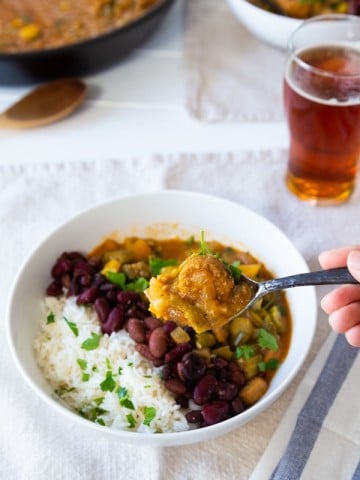 Gumbo in a white bowl with beans and rice with a hand holding a spoon of the stew, and a glass of beer and a skillet of gumbo in the background.