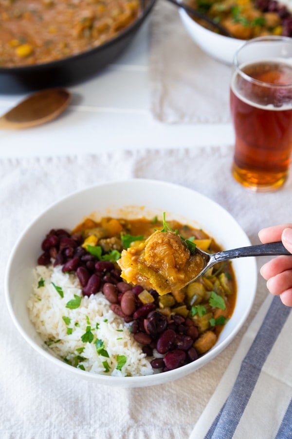 Gumbo in a white bowl with beans and rice with a hand holding a spoon of the stew, and a glass of beer and a skillet of gumbo in the background.