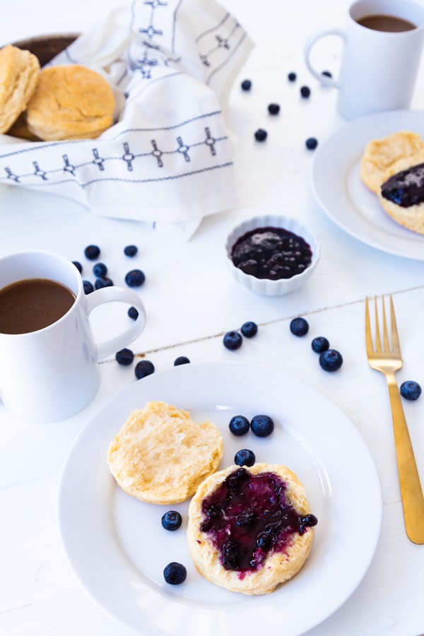 A white plate with a biscuit covered in blueberry jam with a basket of biscuits, a mug of coffee, and a small bowl of blueberries and scattered blueberries on the table
