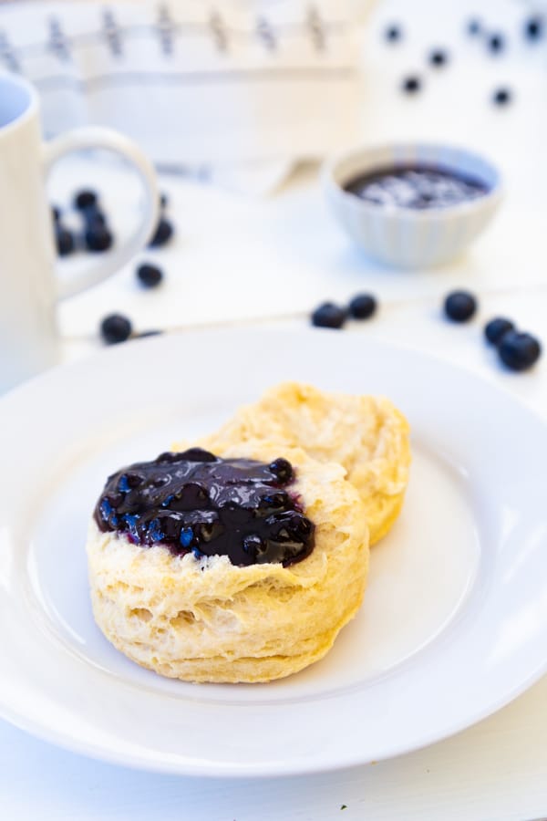 A white plate with a biscuit and blueberry preserves spread on top of it and blueberries on a white table next to the plate