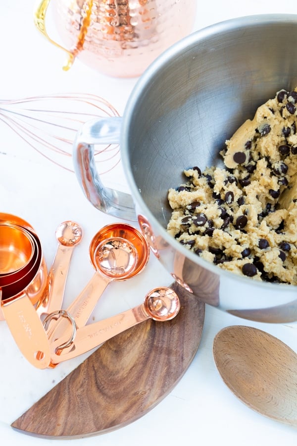 Chocolate chip cookie batter in a silver mixing bowl with copper measuring cups and spoons next to it.