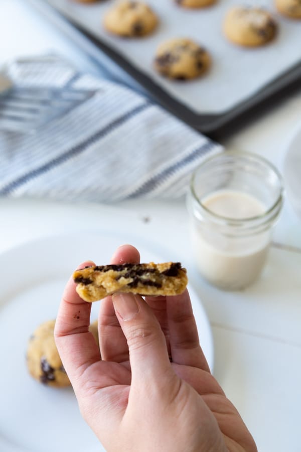 A hand holding half of a chocolate chip cookie and a white plate with another cookie on it, and a glass of milk and a baking sheet with cookies in the background