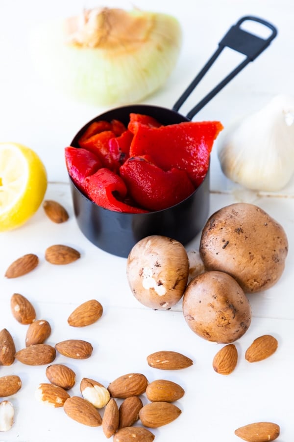 Roasted red peppers in a black measuring cup and mushrooms, almonds, garlic, and lemon on a white table. 