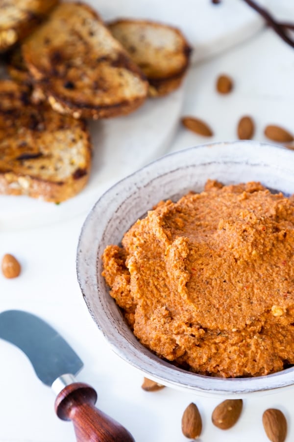 A white bowl of red pepper dip with a wood spread knife and almonds and toast next to the bowl.