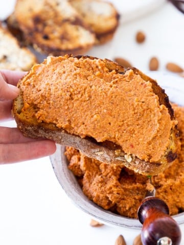 A hand holding a slice of toast with red pepper dip and the bowl of dip and almonds in the background.