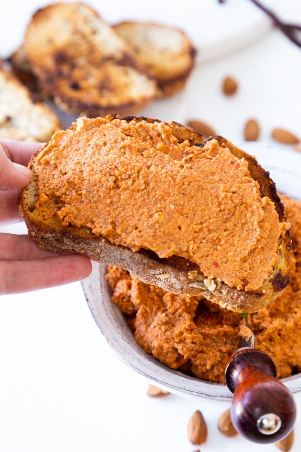 A hand holding a slice of toast with red pepper dip and the bowl of dip and almonds in the background.