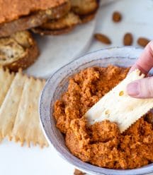 A hand dipping a cracker into a bowl of red pepper pate with crackers and almonds around the bowl on a white table.