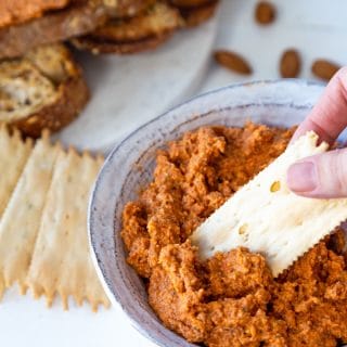 A hand dipping a cracker into a bowl of red pepper pate with crackers and almonds around the bowl on a white table.