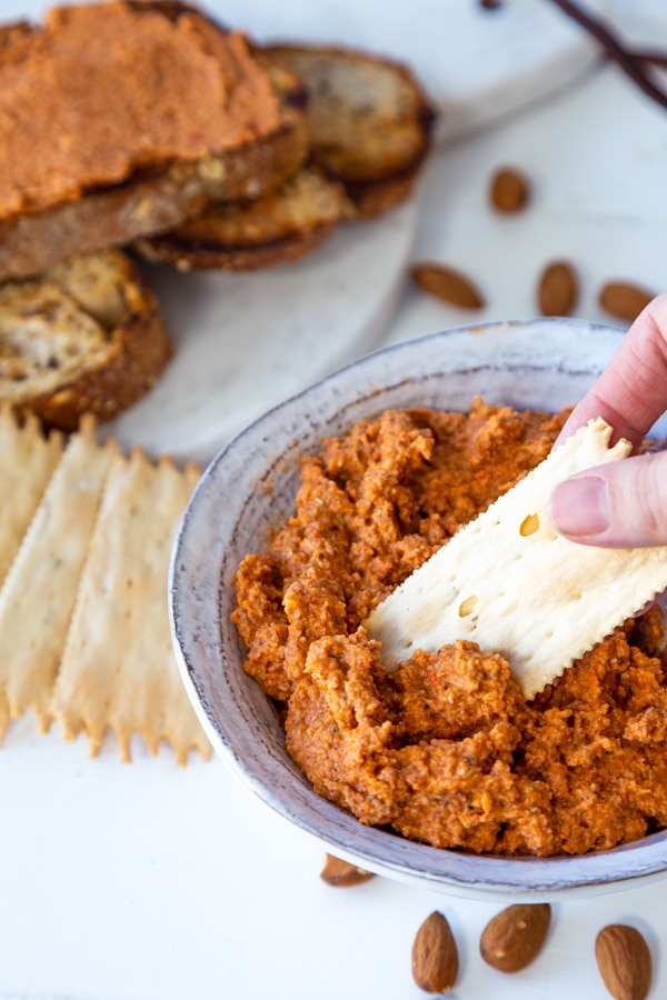 A hand dipping a cracker into a bowl of red pepper pate with crackers and almonds around the bowl on a white table. 
