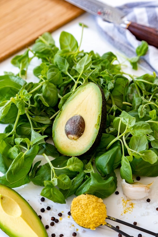 Half of an avocado on top of a pile of basil leaves with lemon, garlic, nutritional yeast, and rock salt and peppercorns.