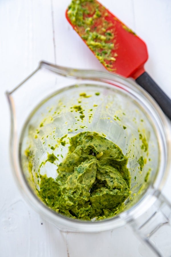 Pesto in a blender with a red and black spatula with oil-free pesto on it on a white board.