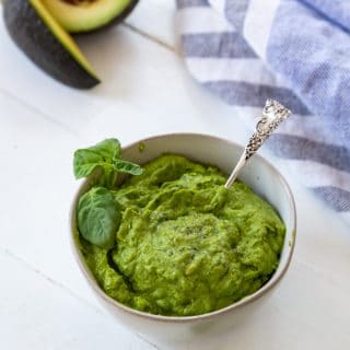 A bowl of pesto with a silver spoon in the pesto and an avocado, basil, and a blue and white towel next to the bowl on a white table.