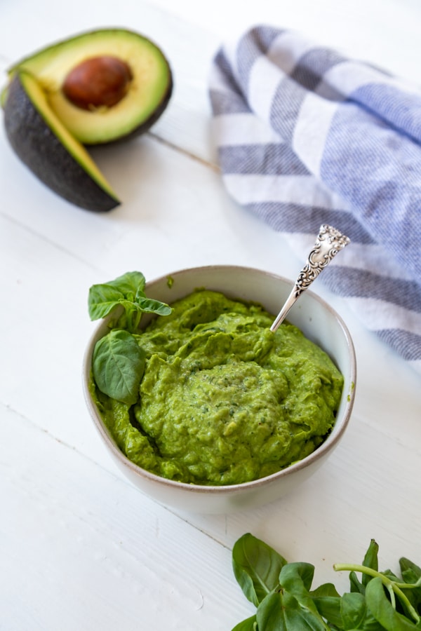 A bowl of oil-free pesto with a silver spoon in the pesto and an avocado, basil, and a blue and white towel next to the bowl on a white table. 