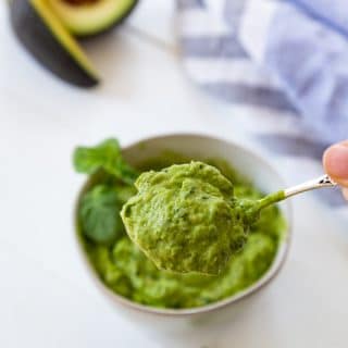 A white bowl with pesto and a hand taking a spoonful out of the bowl. Avocados, basil and a towel are next to the bowl.