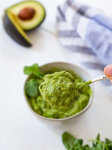 A white bowl with pesto and a hand taking a spoonful out of the bowl. Avocados, basil and a towel are next to the bowl.