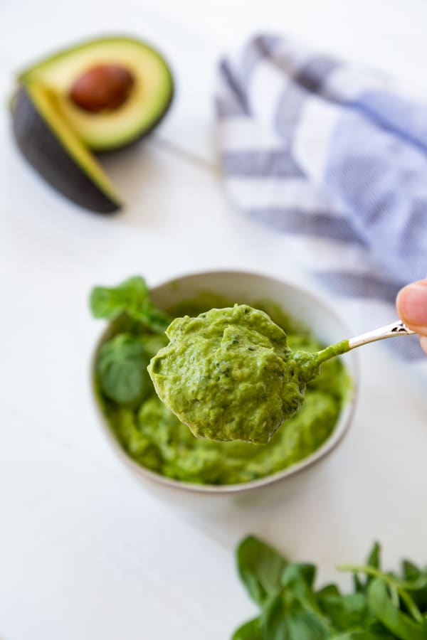 A white bowl with oil-free pesto and a hand taking a spoonful out of the bowl. Avocados, basil and a towel are next to the bowl.