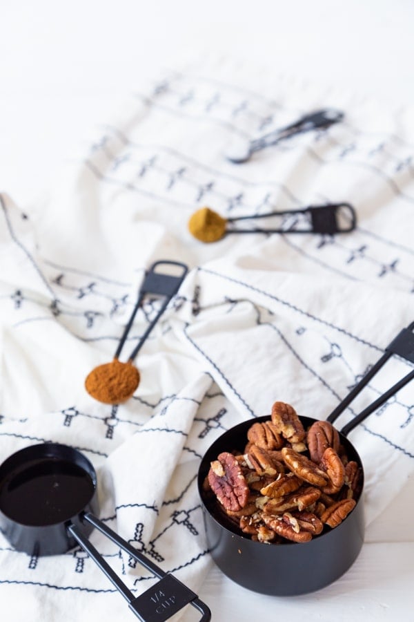 Black measuring cups with pecans, maple syrup, and cumin for spiced pecans