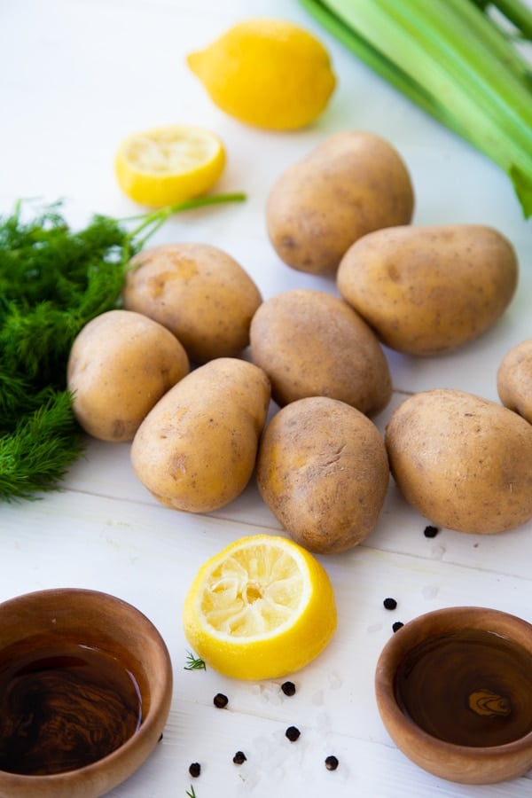 Several whole potatoes, lemon halves, celery, fresh dill and wood bowls with vinegar on a white table.