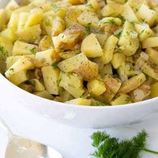 Potato salad in a white bowl with a bunch of dill and a spoon on the side of the bowl.