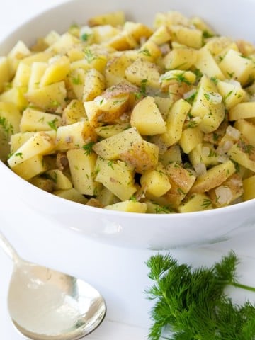 Potato salad in a white bowl with a bunch of dill and a spoon on the side of the bowl.