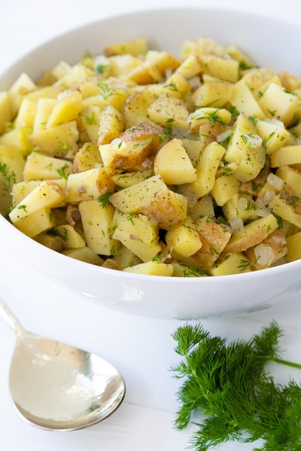 Potato salad in a white bowl with a bunch of dill and a spoon on the side of the bowl.