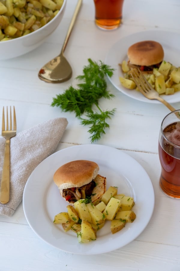 A white wood table with a white plate with a burger and potato salad and a large white bowl of potato salad, glasses of beer, and another plate of food.