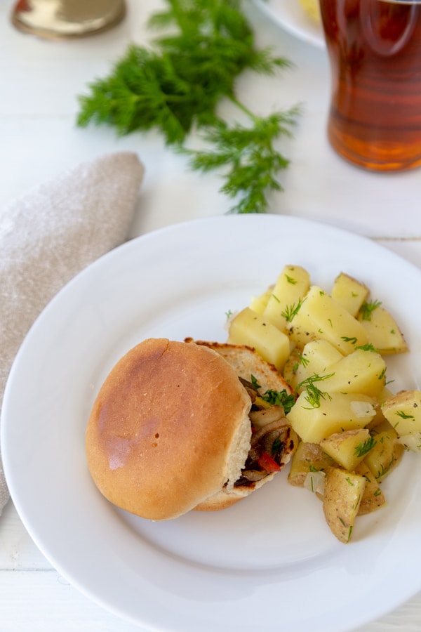 A white plate with a mushroom slider and potato salad, and a glass of beer and fresh dill in the background.