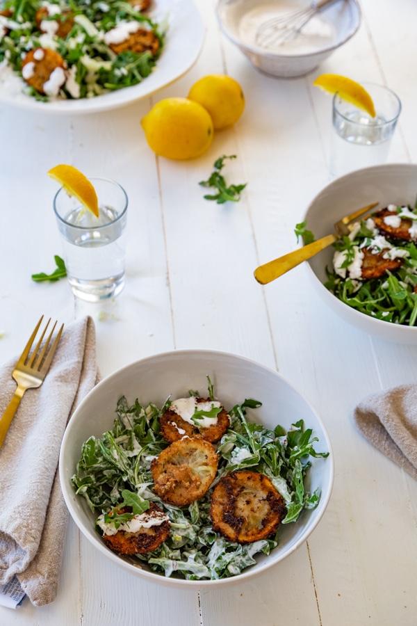 Arugula and fennel salad with fried lemons in three white bowls with salads and lemons on the table and gold utensils.