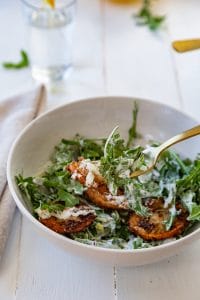 An arugula and fennel salad in a white bowl with a gold fork and a glass of water with lemon in the background and fried lemons by Veganosity.