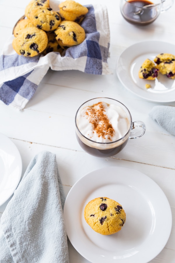 A white plate with one blueberry muffin and a cup of coffee and basket of muffins in the background. 