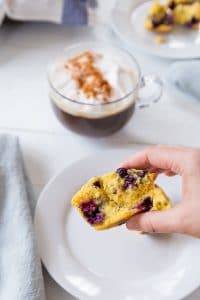 A hand holding a blueberry muffin over a white plate with a mug of coffee and another white plate with muffins in the background.