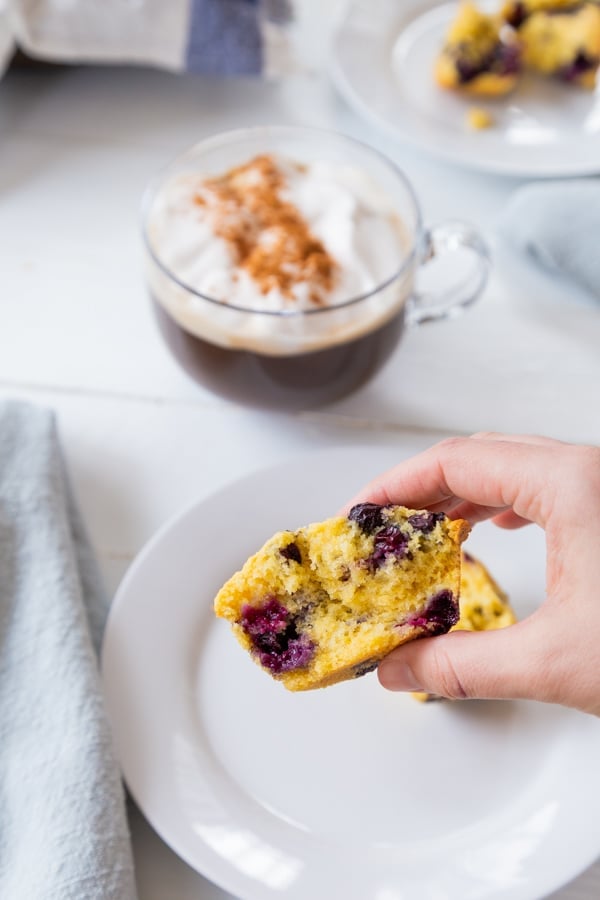 A hand holding a blueberry muffin over a white plate with a mug of coffee and another white plate with muffins in the background. 