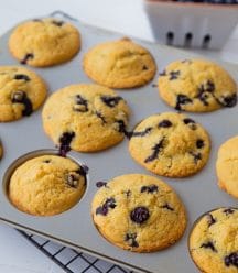 Blueberry muffins in a muffin tin with a white ceramic dish of blueberries in the background.