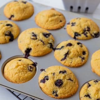 Blueberry muffins in a muffin tin with a white ceramic dish of blueberries in the background.