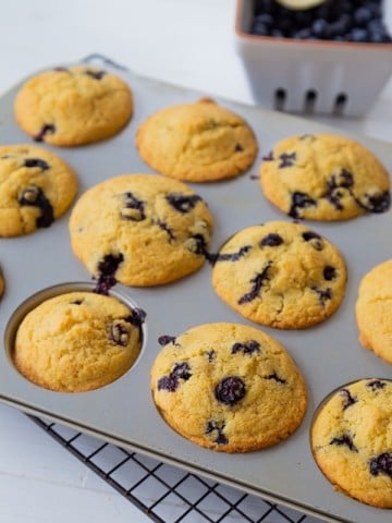 Blueberry muffins in a muffin tin with a white ceramic dish of blueberries in the background.