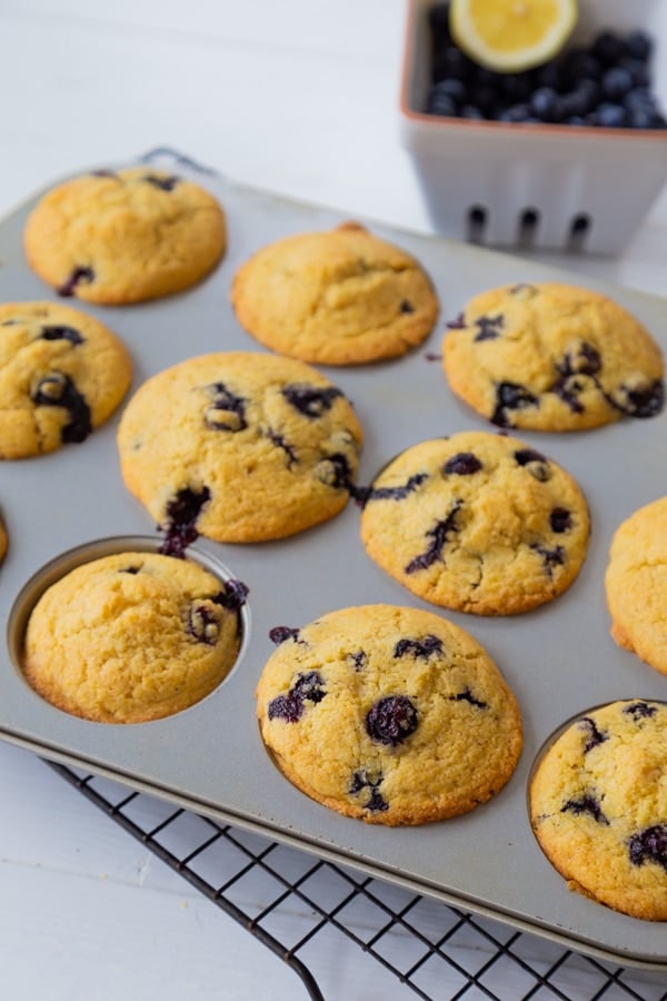 Blueberry muffins in a muffin tin with a white ceramic dish of blueberries in the background.