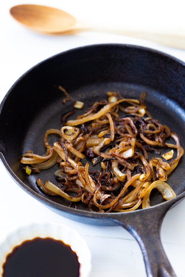 Fried onions in an iron skillet with a white dish of soy sauce and a wood spoon next to it. 