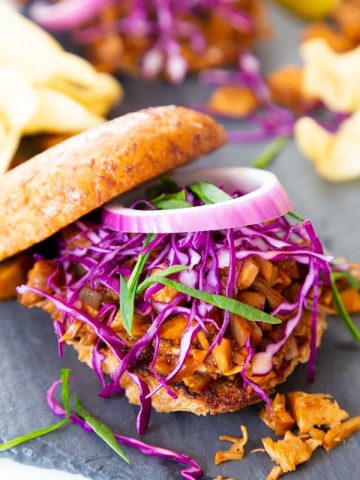 A pulled BBQ sandwich featured in the Veganosity cookbook Great Vegan BBQ Without a Grill with the bun half on the top and onions and slaw on top of the sandwich