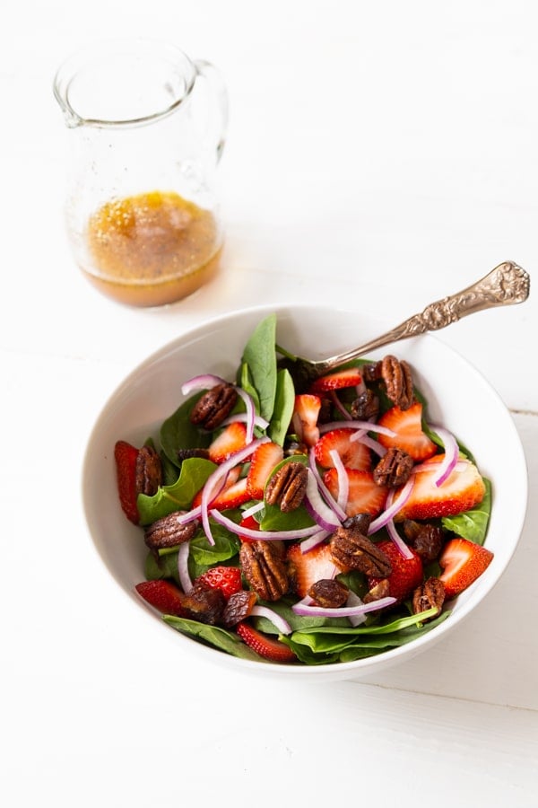 A salad with strawberries and pecans in a white bowl with salad dressing next to it