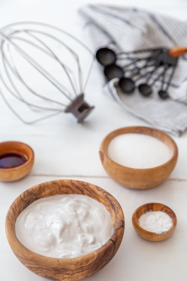 Wood bowls filled with coconut milk, sugar, and vanilla extract with a wire whisk and black measuring spoons on a white wood table. 